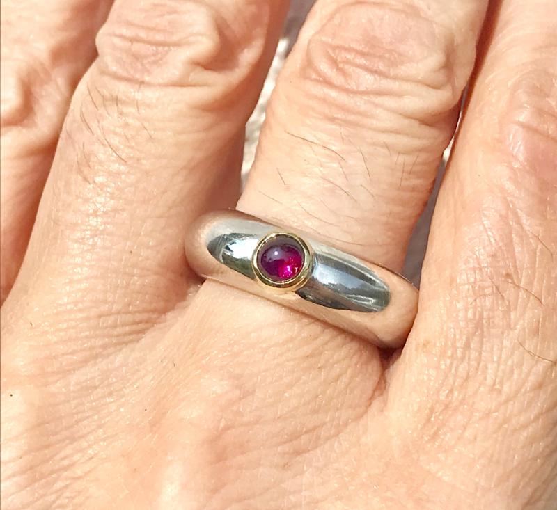 Ein runder rosaner Rhodolithcabochon ist in einer Goldfassung aus 750er Gold gefasst und sitzt auf einer kräftigen und aus einem Stück gegossenen Ringschiene aus 935er Silber.